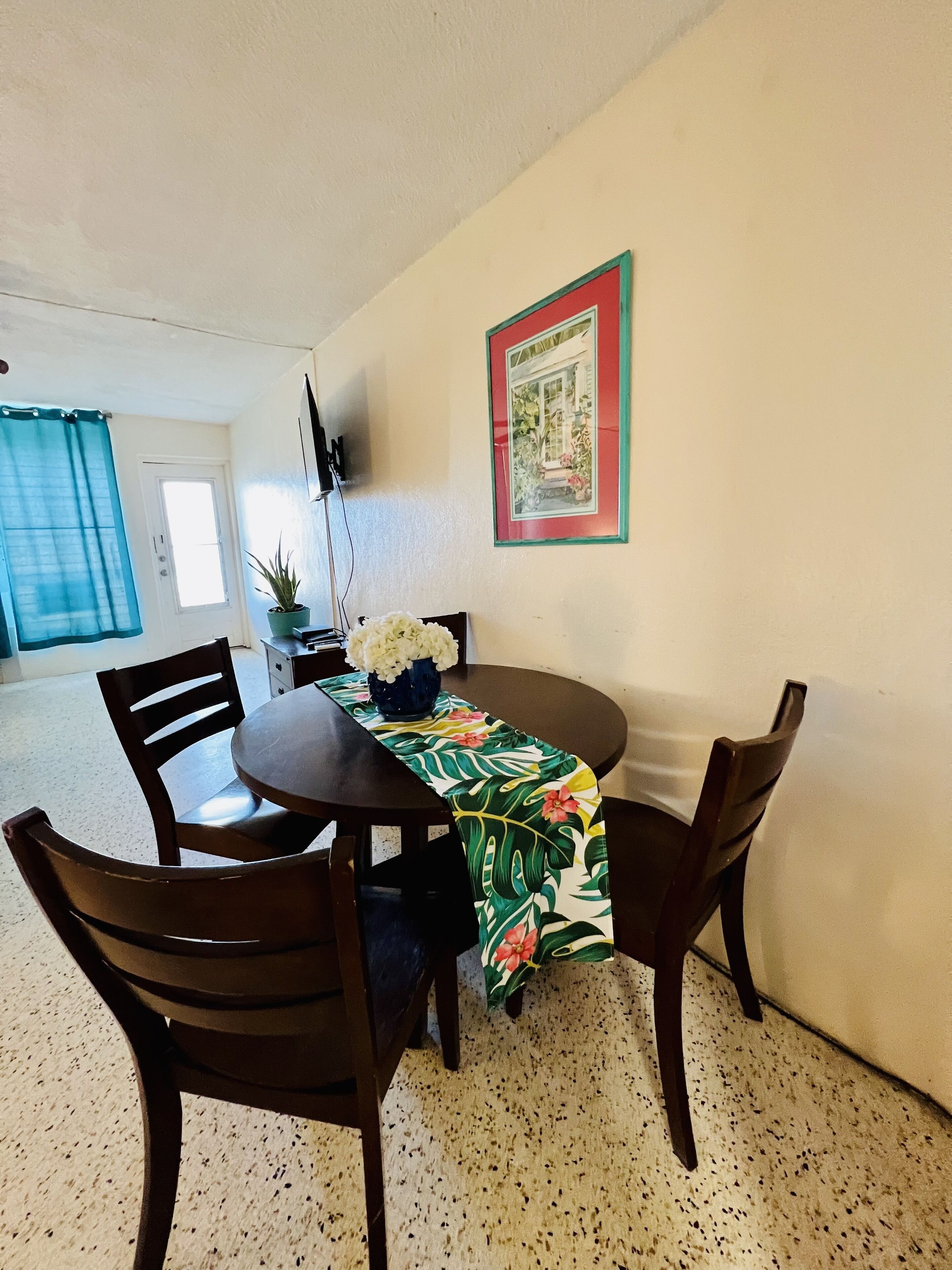 A dining area with a round table, four chairs, a floral table runner, a potted plant, and a framed picture on the wall.