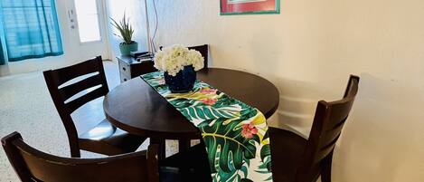 A dining area with a round table, four chairs, a floral table runner, a potted plant, and a framed picture on the wall.