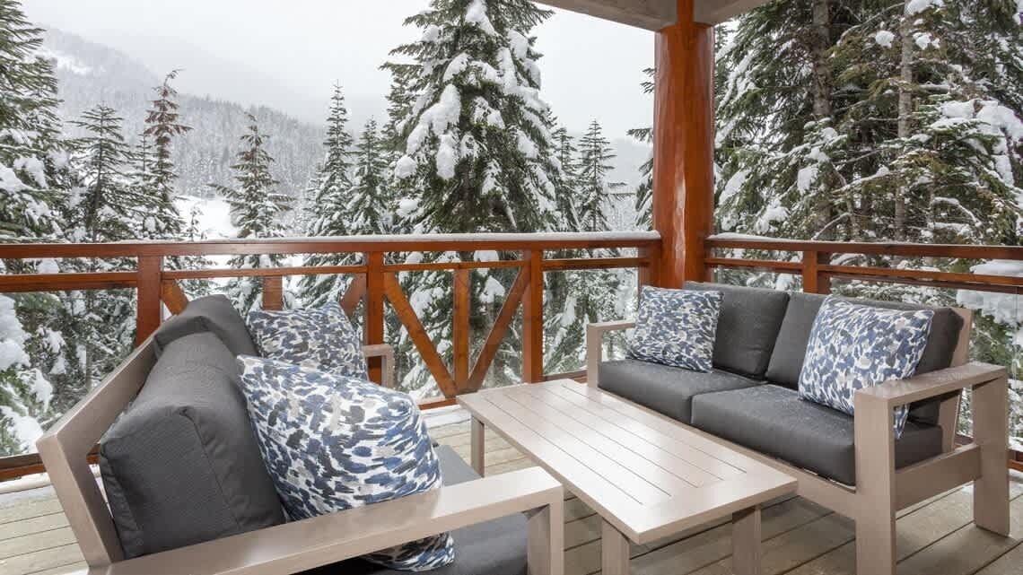 Een houten terras met een bank en een tafel, met uitzicht op een besneeuwd bos.