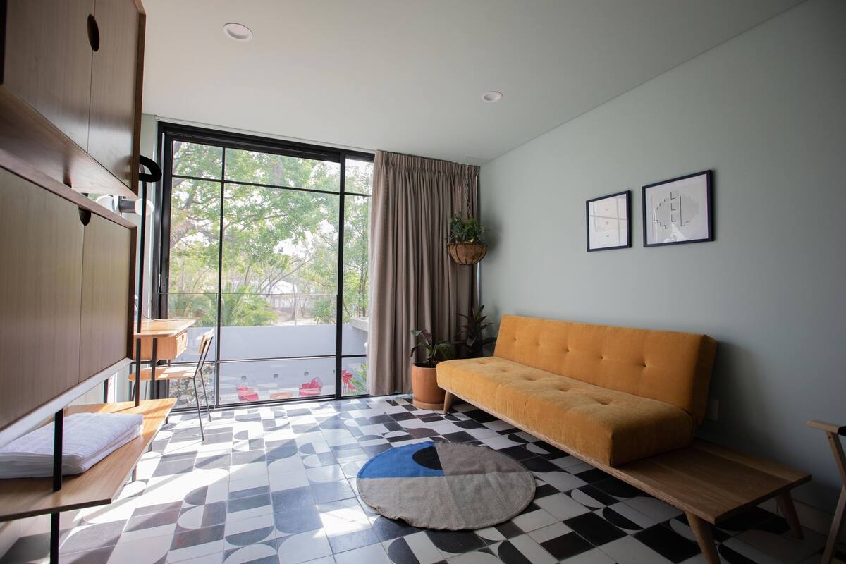 Una sala de estar moderna con un sofá, una mesa de comedor y sillas. Hay un ventanal con cortinas, una planta en maceta y dos cuadros en la pared.