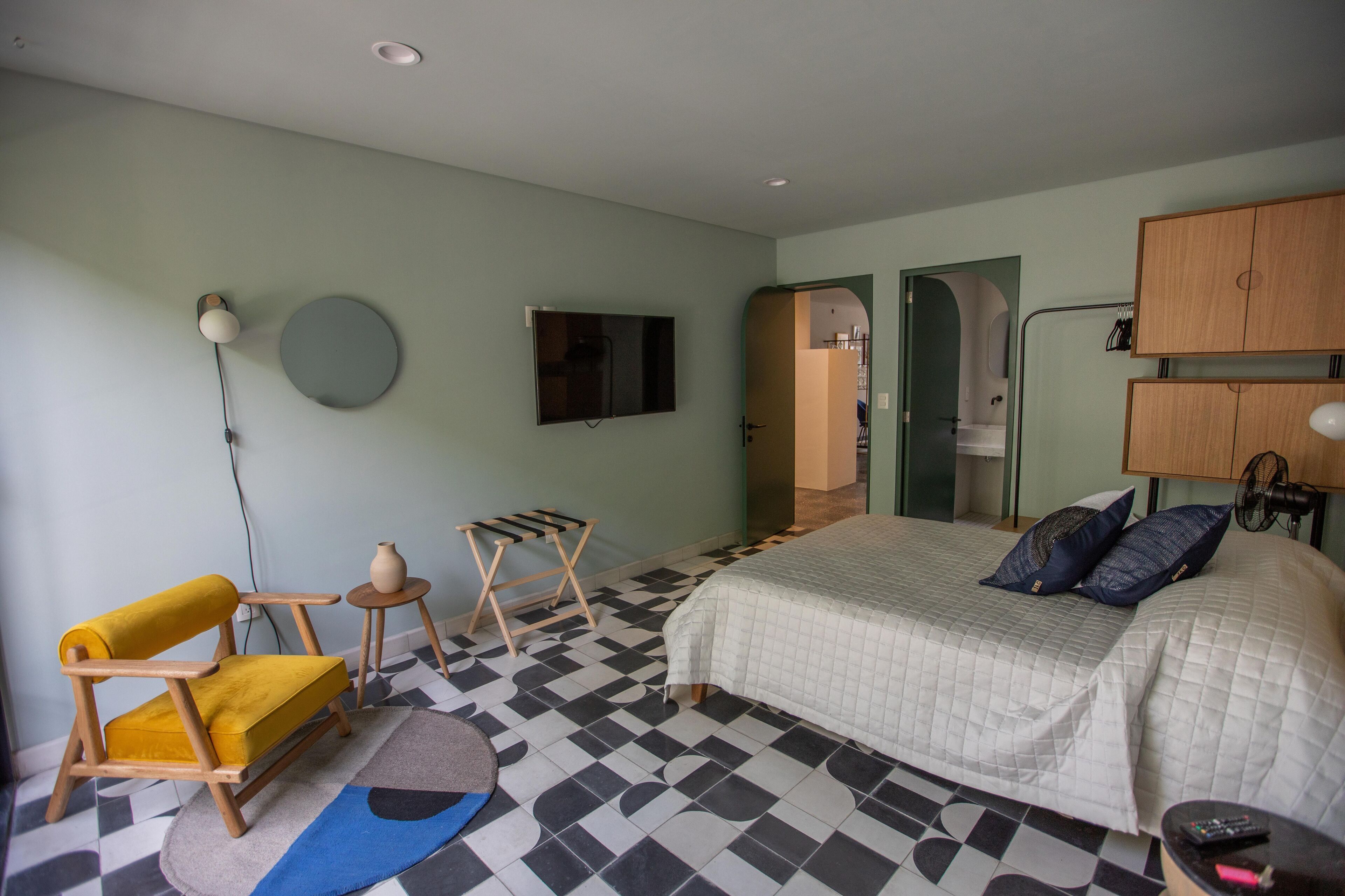 A bedroom with a bed, a chair, a small table, a mirror, and a television on the wall.