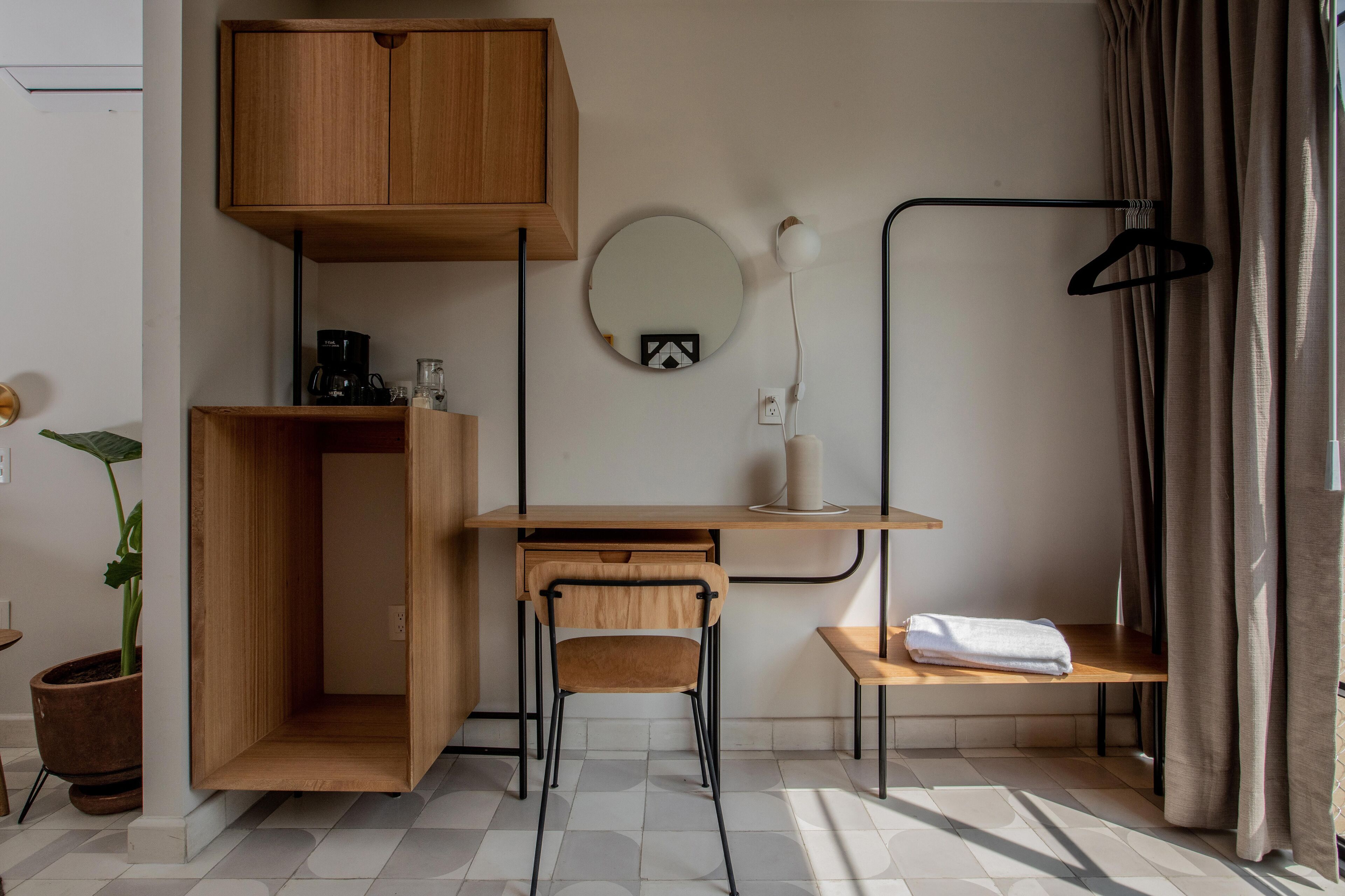 A compact room with a wooden desk, a chair, a hanging light, a mirror, and a towel on a small table.