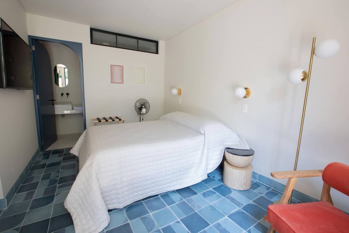 Un dormitorio con cama, una silla, un espejo, un ventilador y un lavabo.