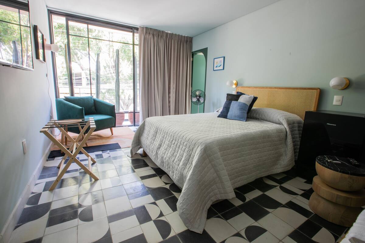 Una habitación de hotel con cama, sillón, televisión y ventana con cortinas.