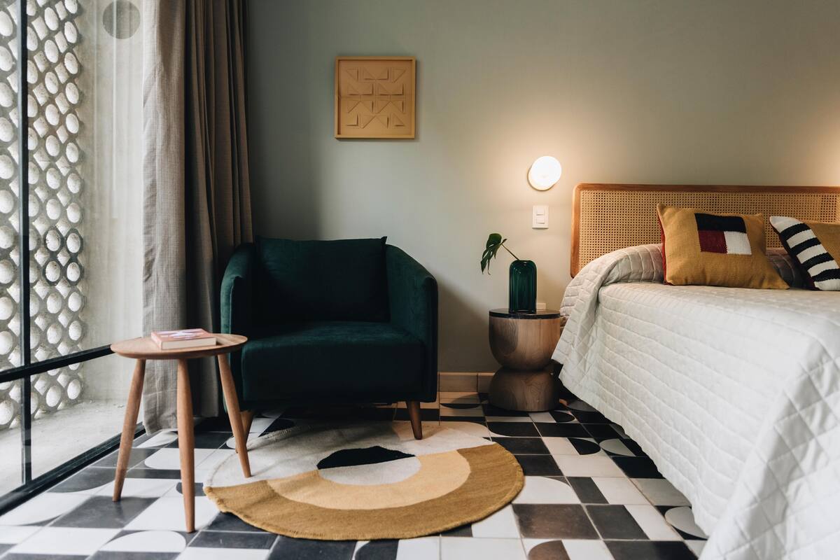 Un dormitorio con una cama, un sillón verde, una mesita redonda pequeña, una planta y un suelo con estampado geométrico.
