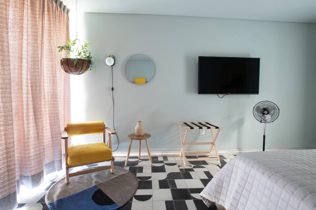 Una habitación de hotel moderna con una cama, un sillón amarillo, una mesita con un florero, un espejo redondo, un televisor de pantalla plana y un ventilador de pie.