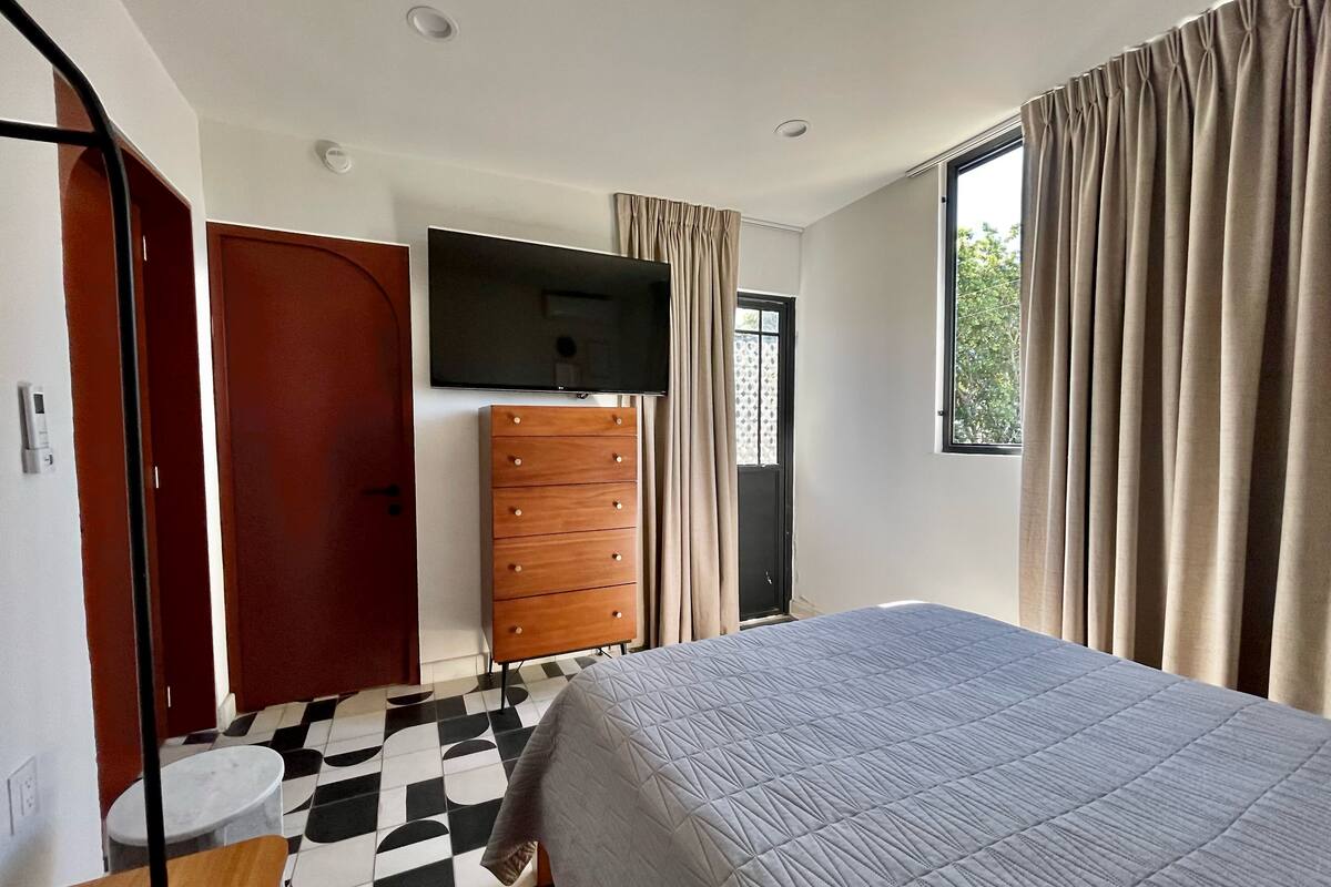 Un dormitorio con cama, televisor montado en la pared, cómoda, ventana con cortinas y suelo estampado.