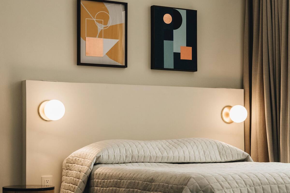 Una cama bien hecha con una colcha acolchada, dos lámparas de pared y dos cuadros abstractos en la pared.