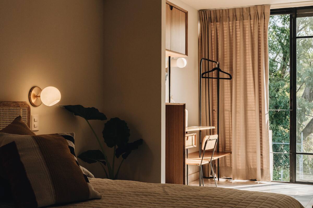 Un dormitorio con cama, lámpara de noche, planta colgante y vistas a la vegetación por la ventana.