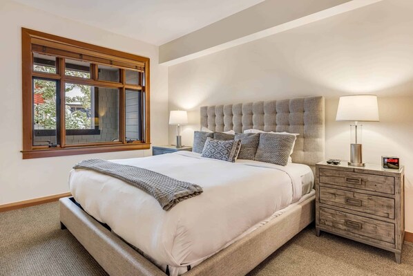 Un dormitorio con una cama grande, dos mesitas de noche con lámparas, un cabecero de madera y una ventana con vistas a árboles.