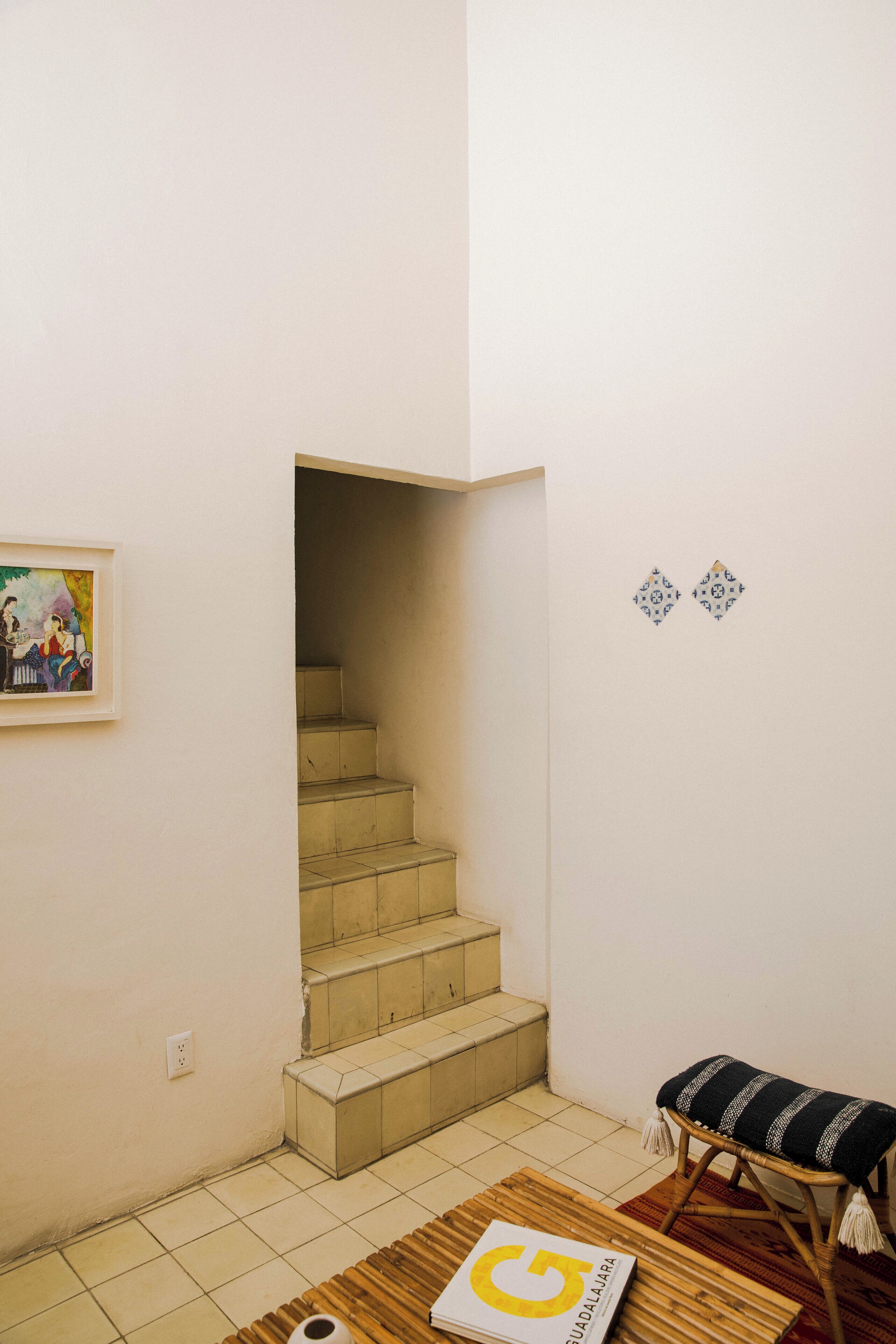 Una escalera que lleva a un piso superior, un cuadro enmarcado en la pared, un banco con cojín y un libro en el suelo.