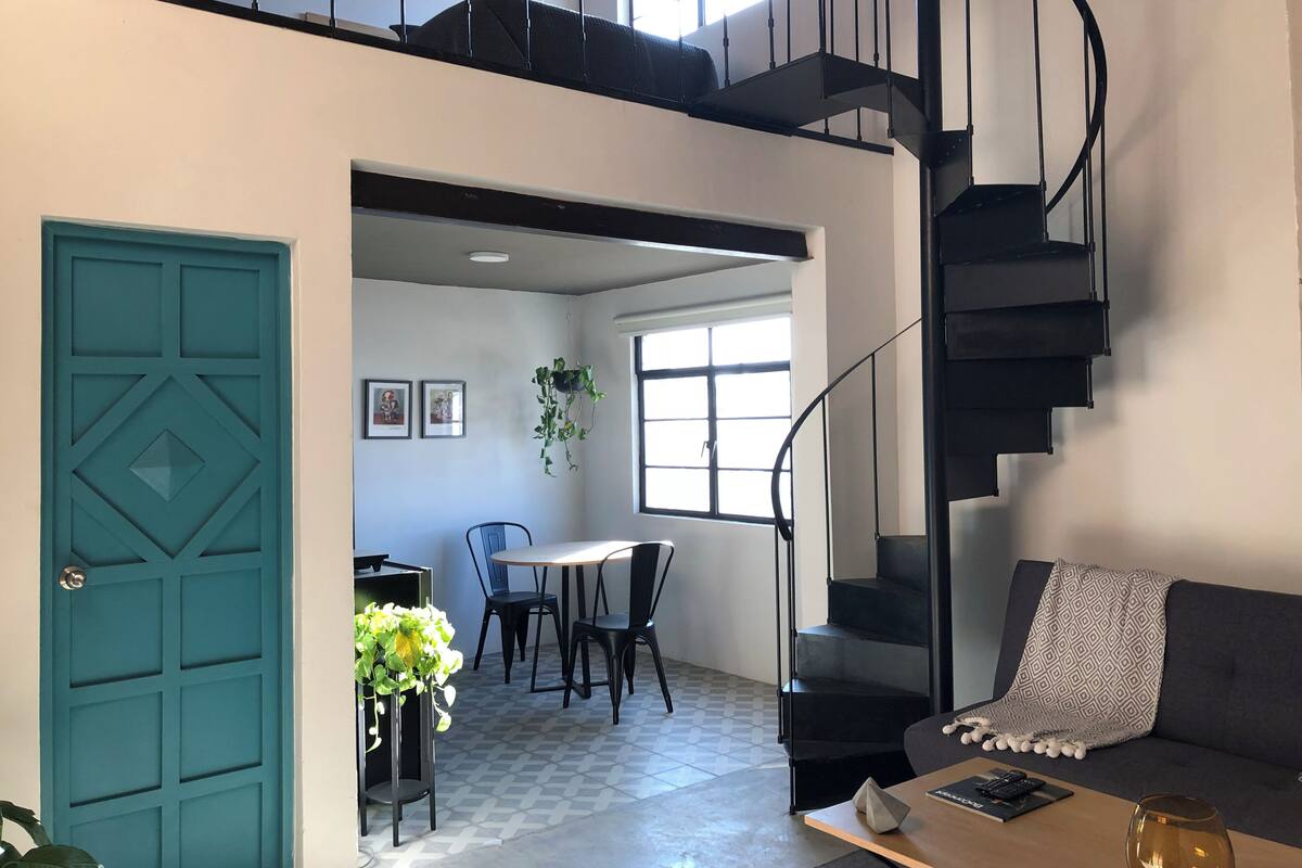 Un intérieur moderne avec une porte de couleur turquoise, un escalier, un coin repas avec des chaises et une table, et un canapé recouvert d’une couverture.
