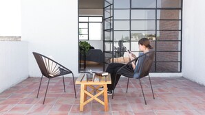 A patio with two black wireframe chairs, a wooden table, and a person using a laptop.