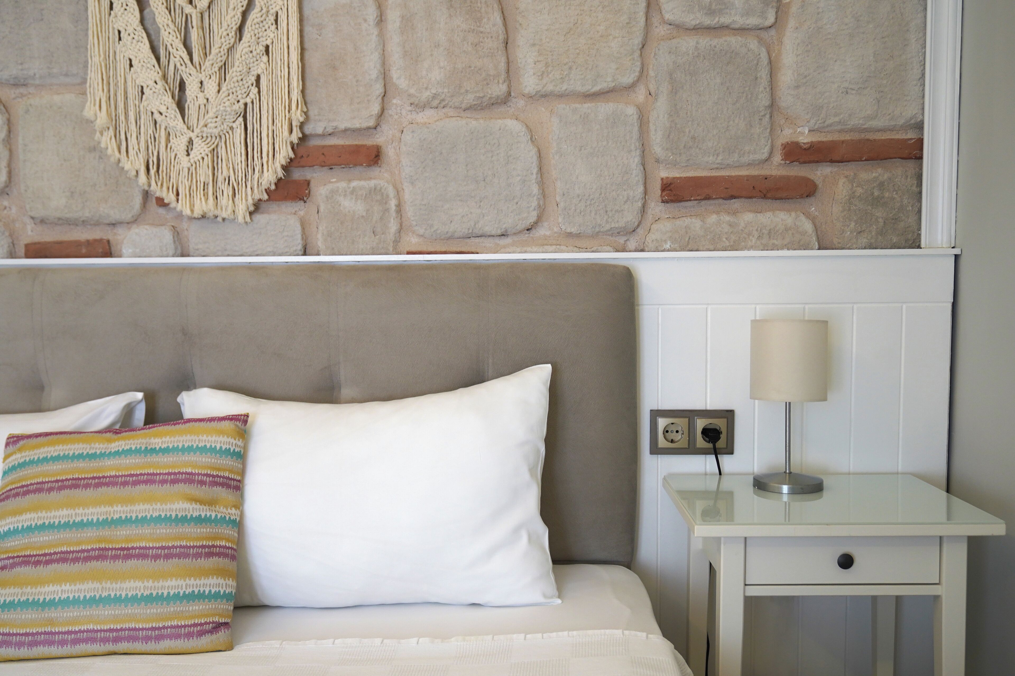 Een net opgemaakt bed met een wit kussen en een kleurrijk gestreept kussen, een nachtkastje met een lamp, en een stenen muur op de achtergrond.