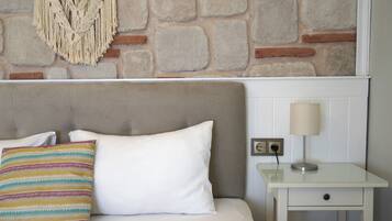 Una cama bien tendida con un almohadón blanco y uno colorido a rayas, una mesita de noche con una lámpara, y una pared de piedra al fondo.