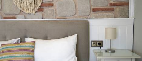 Een net opgemaakt bed met een wit kussen en een kleurrijk gestreept kussen, een nachtkastje met een lamp, en een stenen muur op de achtergrond.