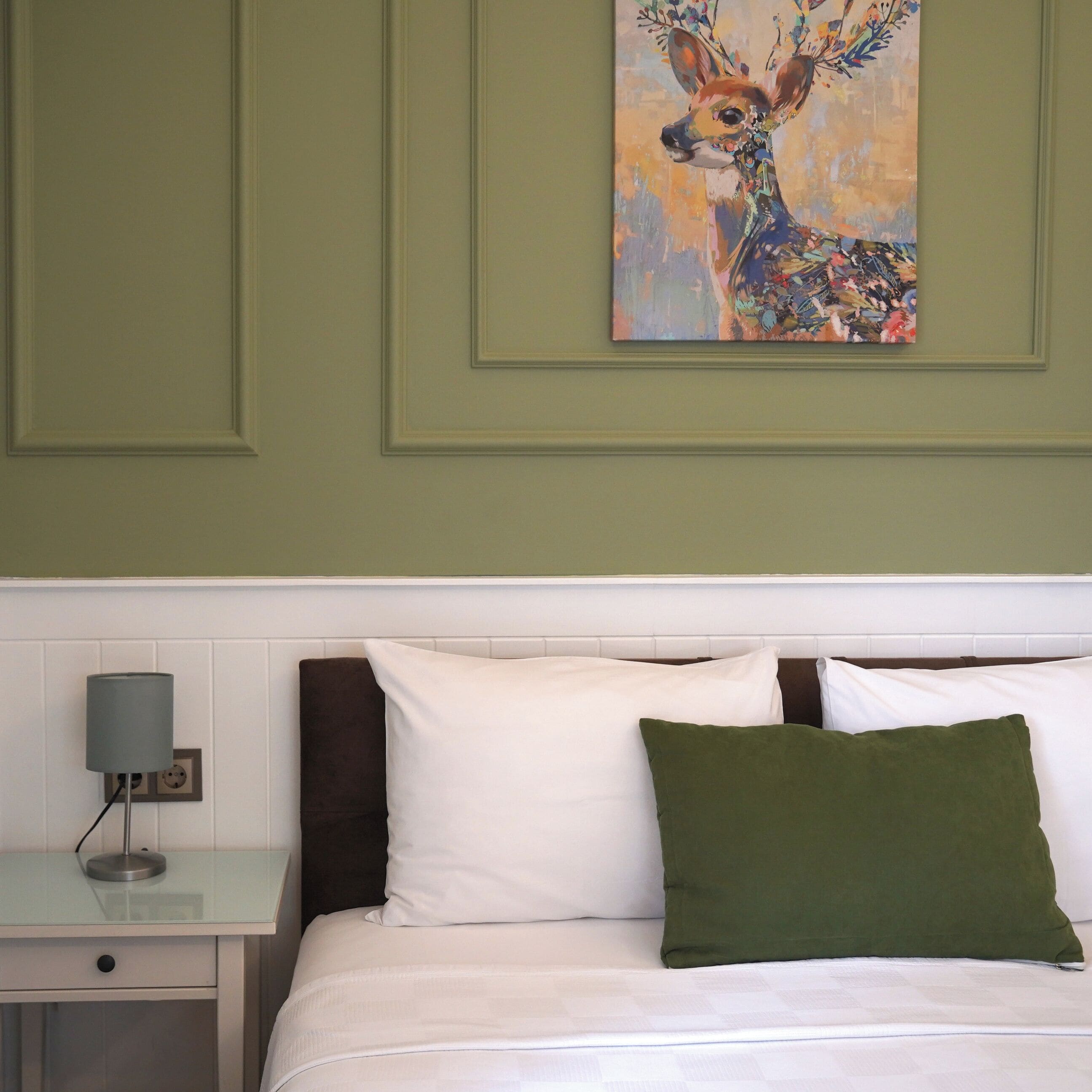 Una cama bien hecha con almohadas blancas y verdes, una lámpara de noche y un cuadro de un ciervo en la pared.