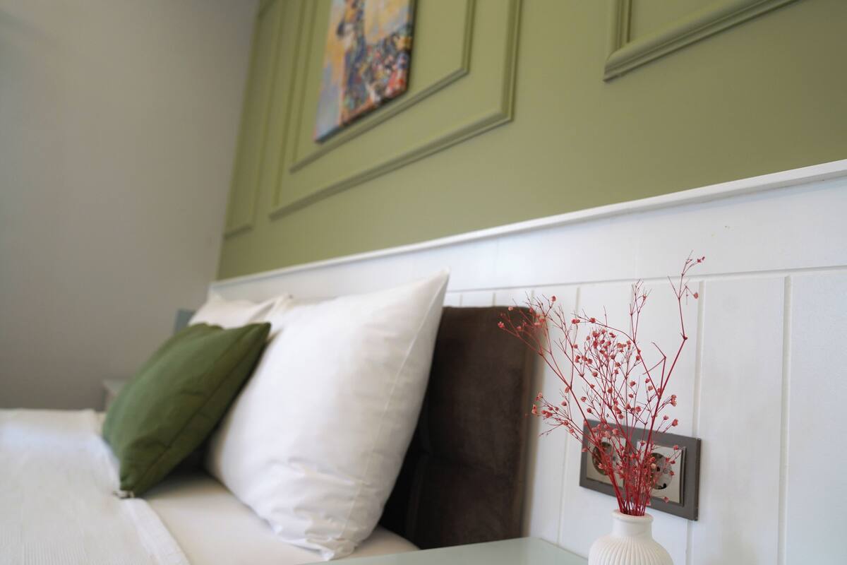 Une chambre à coucher avec un lit, des oreillers, un vase contenant des branches rouges et un tableau accroché au mur.