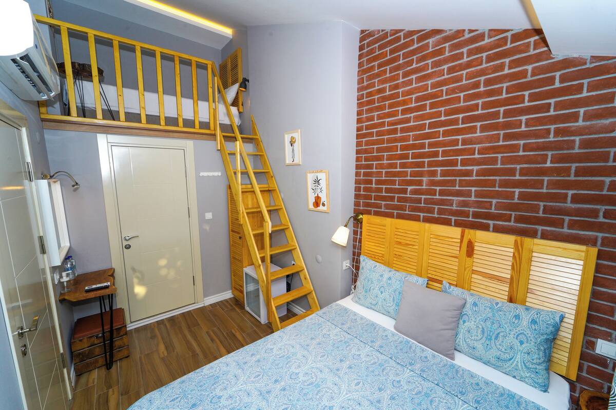 Une chambre avec un lit en mezzanine, une échelle en bois, un mur en briques et une petite table avec une lampe.