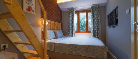 A bedroom with a bed, a wooden staircase, a window with curtains, and a television mounted on the wall.