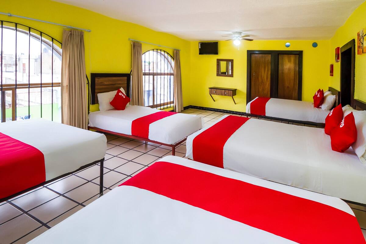 A hotel room with four beds, each with red and white bedding. There is a window with curtains, a television on the wall, and a wooden door.