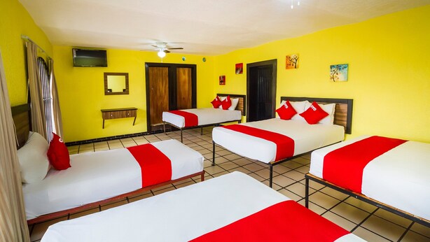 Quarto de hotel com quatro camas, cada uma com roupa de cama branca e vermelha, um ventilador de teto, televisão e obras de arte nas paredes.