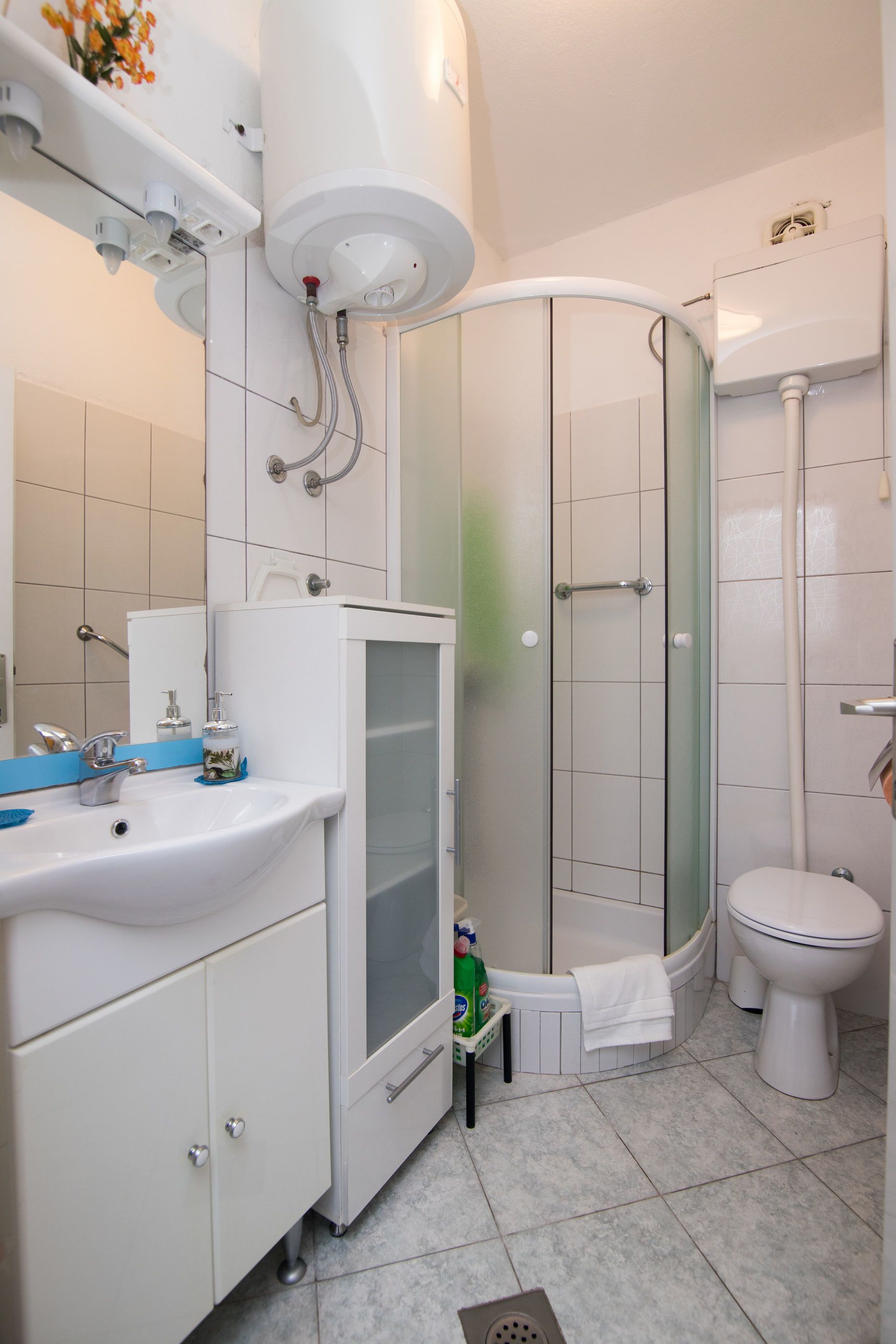 Et moderne badeværelse med en hvid vask, spejl og toilet, samt en brusekabine med glasdøre og et hvidt skab med en lille trinbræt.