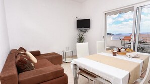 Une pièce moderne avec un canapé marron, une table à manger recouverte d’une nappe blanche, un téléviseur fixé au mur et une vue sur la mer par de grandes fenêtres.