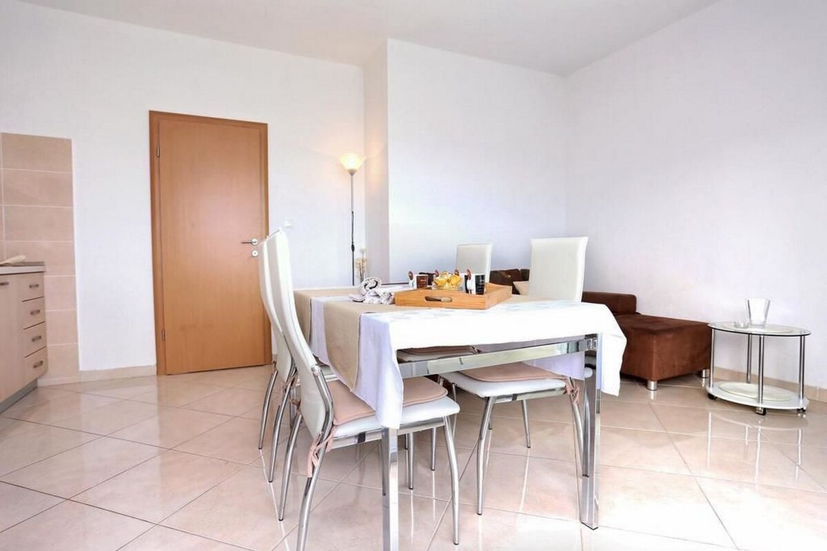 A modern dining area with a table set for two, a chair, a small side table, and a door in the background.