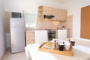 A compact kitchen with a refrigerator, stove, oven, and wooden cabinets. A breakfast tray with bread, coffee cups, and a teapot is on the table. - Apartments Frano (Seget Vranjica)