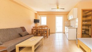 Una sala de estar con un sofá, una mesa de madera y un televisor. Se observa una cocina con un fregadero y armarios.