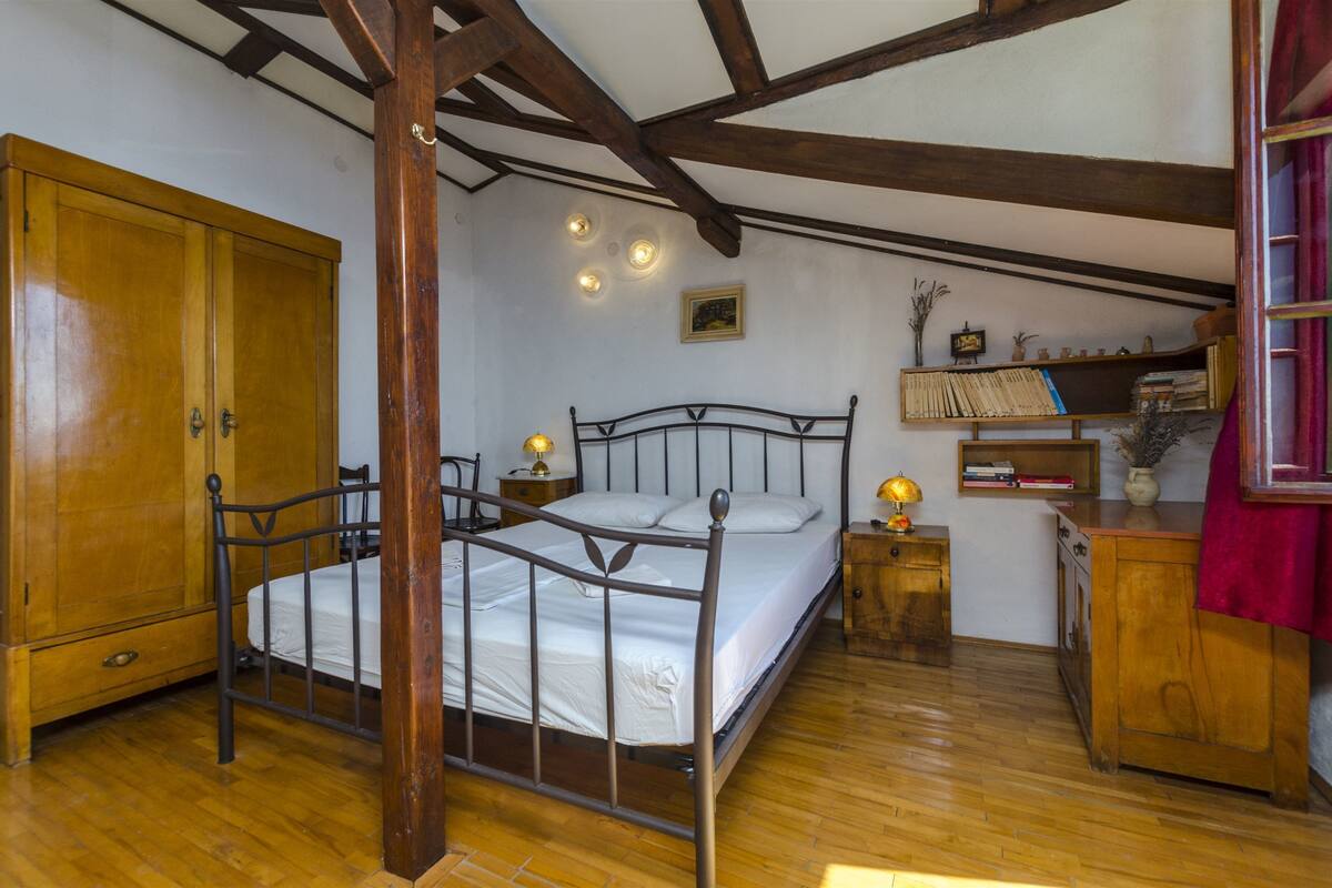 Un dormitorio con un armario de madera, estructura de cama metálica, mesita de noche con lámpara y estante con libros y objetos decorativos.