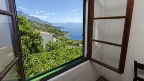 A room with a large window offering a view of a coastal landscape with mountains and a body of water. - Apartment Boris (Brela)