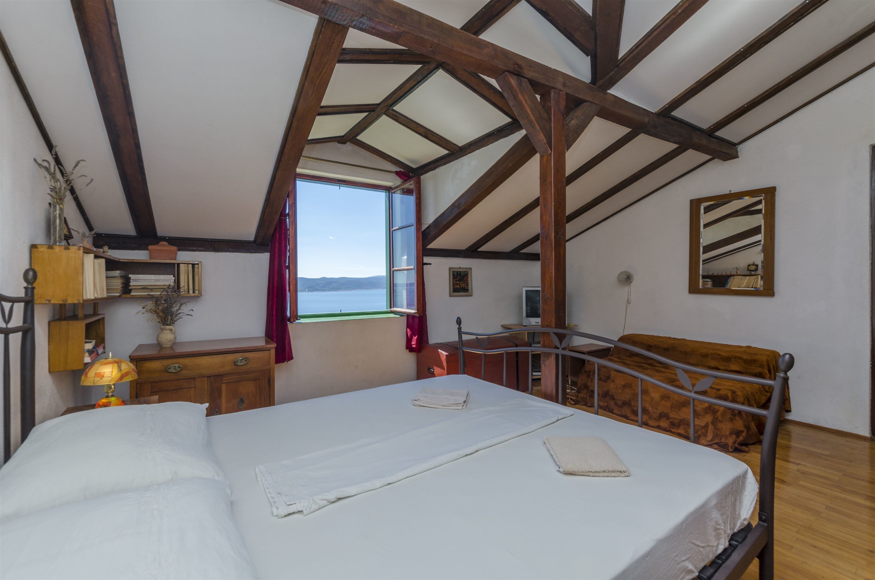 A bedroom with a bed, a wooden ceiling, a window with a view, a bedside table, and a mirror on the wall.