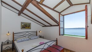 A bedroom with a wooden ceiling, a bed with a red bench, a nightstand, a lamp, and a window with a view of the sea. - Apartment Boris (Brela)