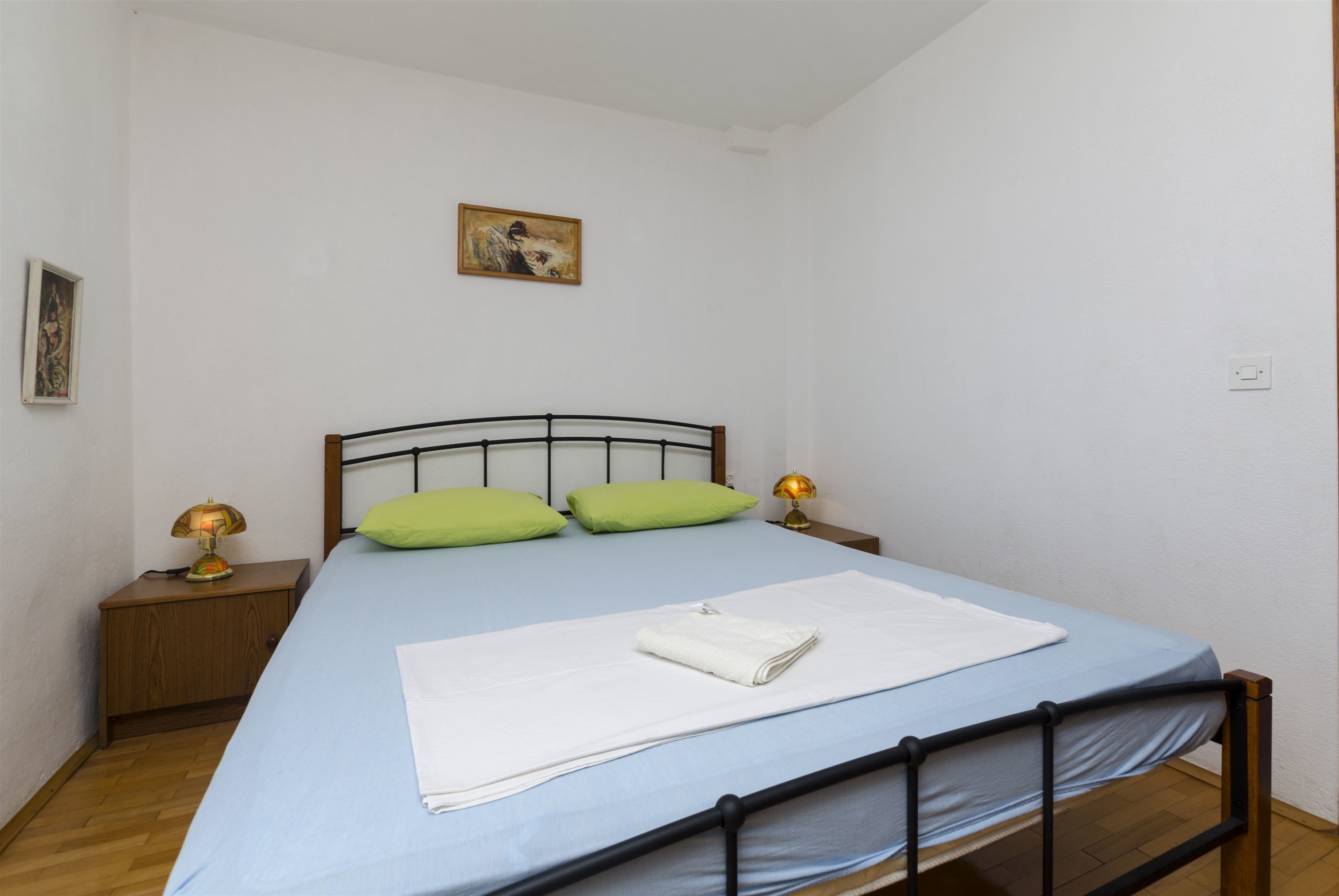 Ein ordentlich bezogenes Bett mit einer blauen Bettdecke, zwei grünen Kissen und einem weißen Handtuch. Ein kleiner Nachttisch mit einer Lampe steht daneben.
