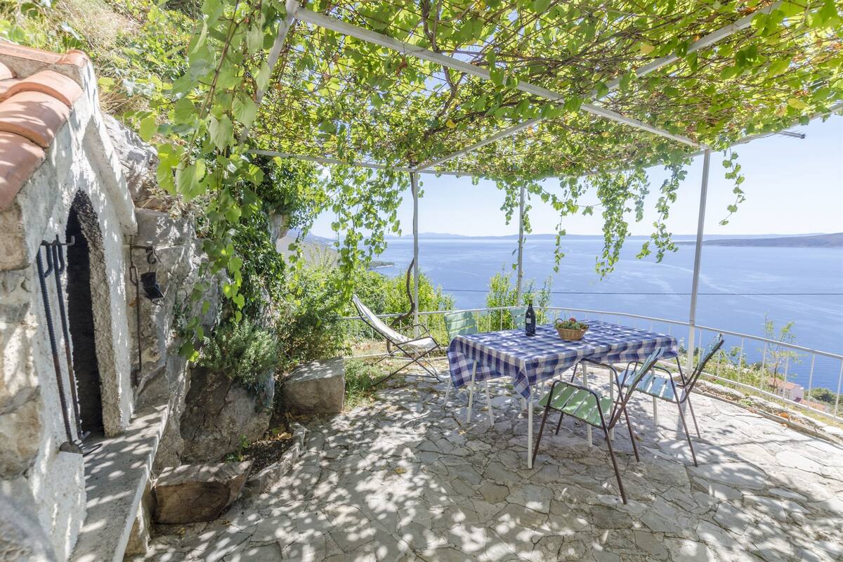 Un patio de piedra con una mesa y sillas bajo una pérgola cubierta de hiedra, con vista a un cuerpo de agua.