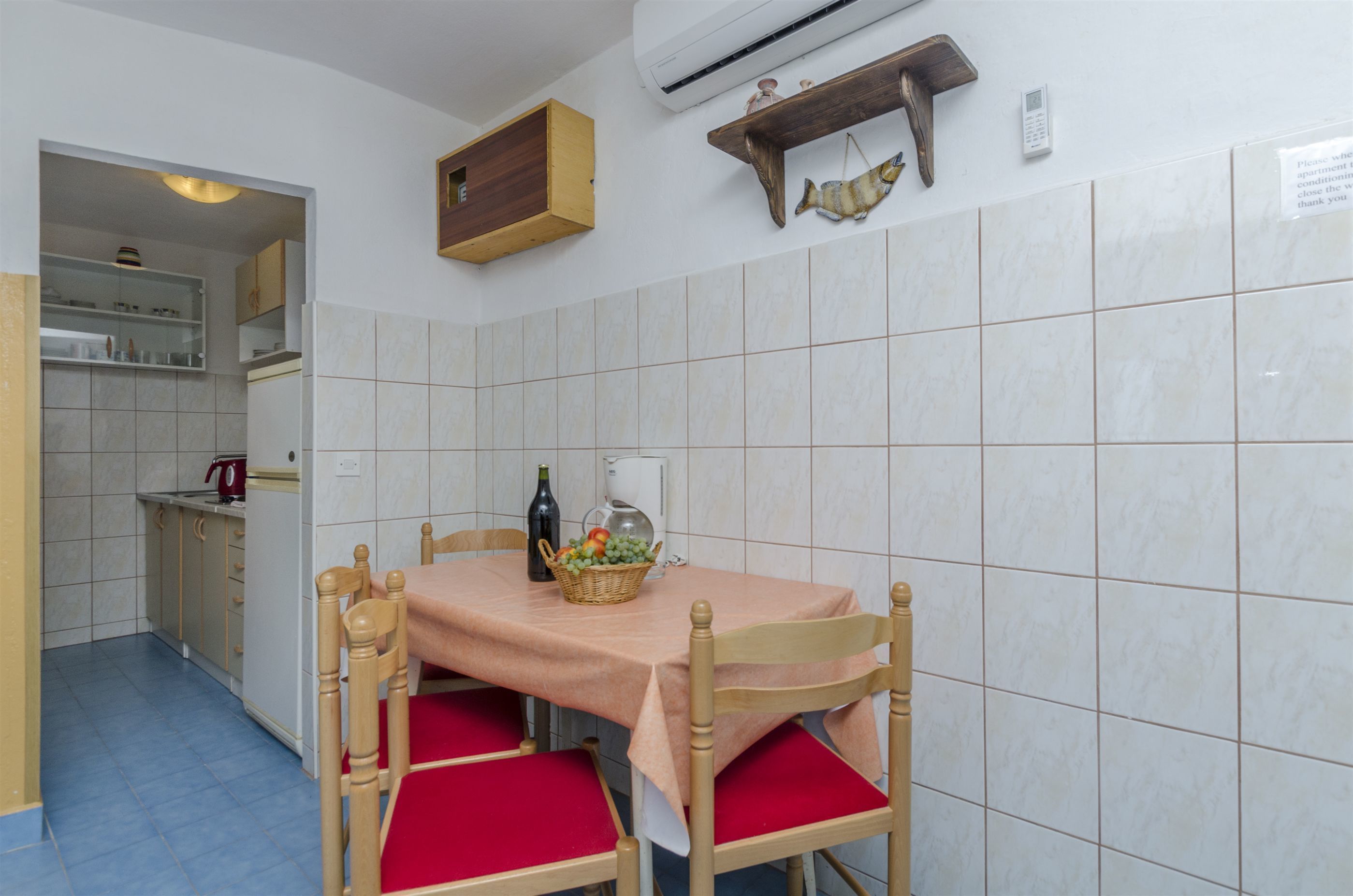 A dining area with a table set for four, a kitchen in the background, and a wall-mounted shelf with decorative items.