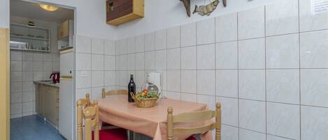 A dining area with a table set for four, a kitchen in the background, and a wall-mounted shelf with decorative items.