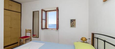 A bedroom with a bed, a wooden wardrobe, a chair, a window with a view, and a small table with a lamp.