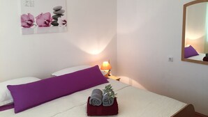 Cama bem arrumada com almofadas roxas, uma toalha enrolada e uma pequena planta na mesinha de cabeceira.