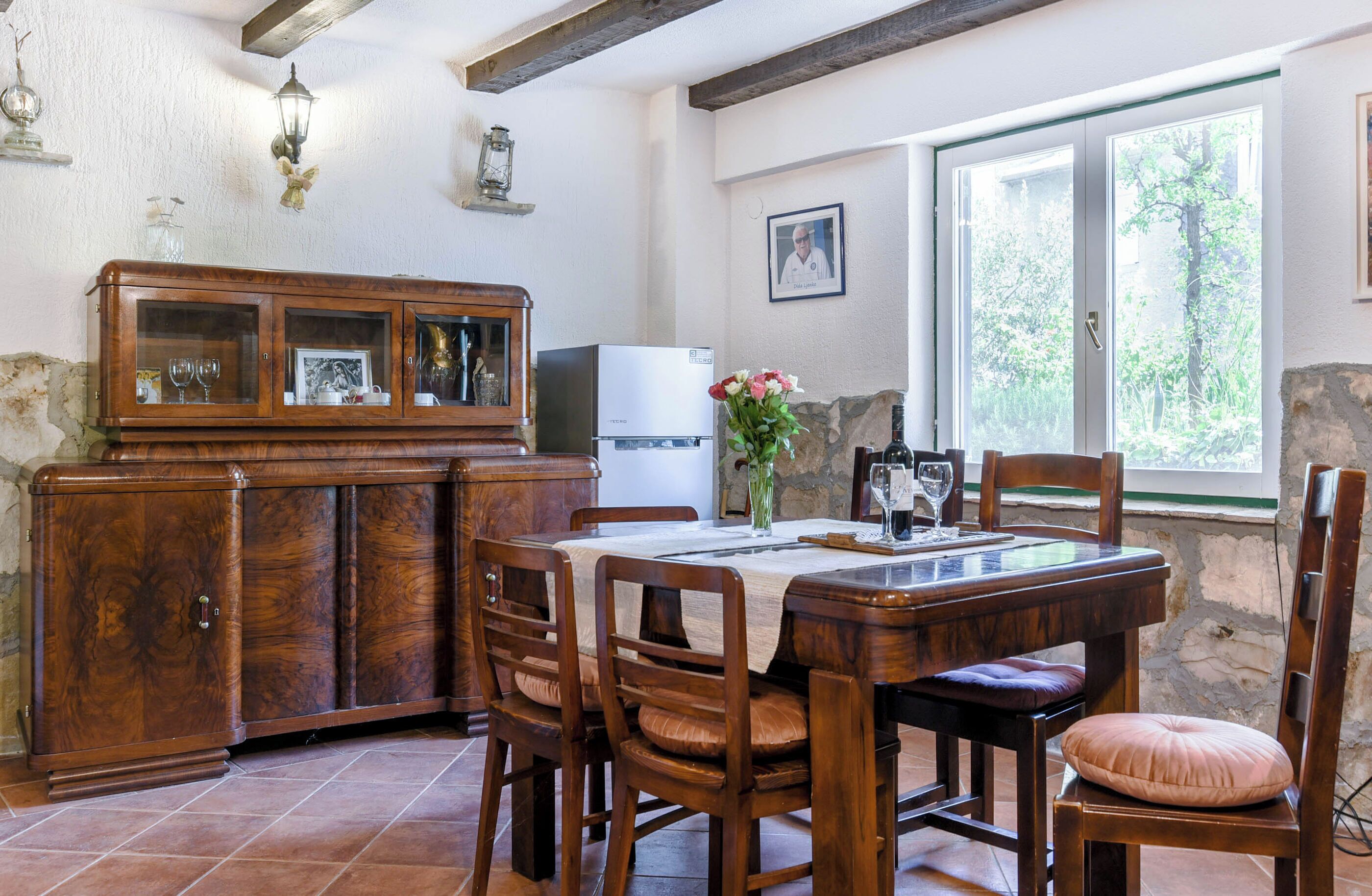 Un coin repas avec une table et des chaises en bois, un meuble avec des portes en verre, un réfrigérateur et une fenêtre donnant sur un paysage verdoyant.