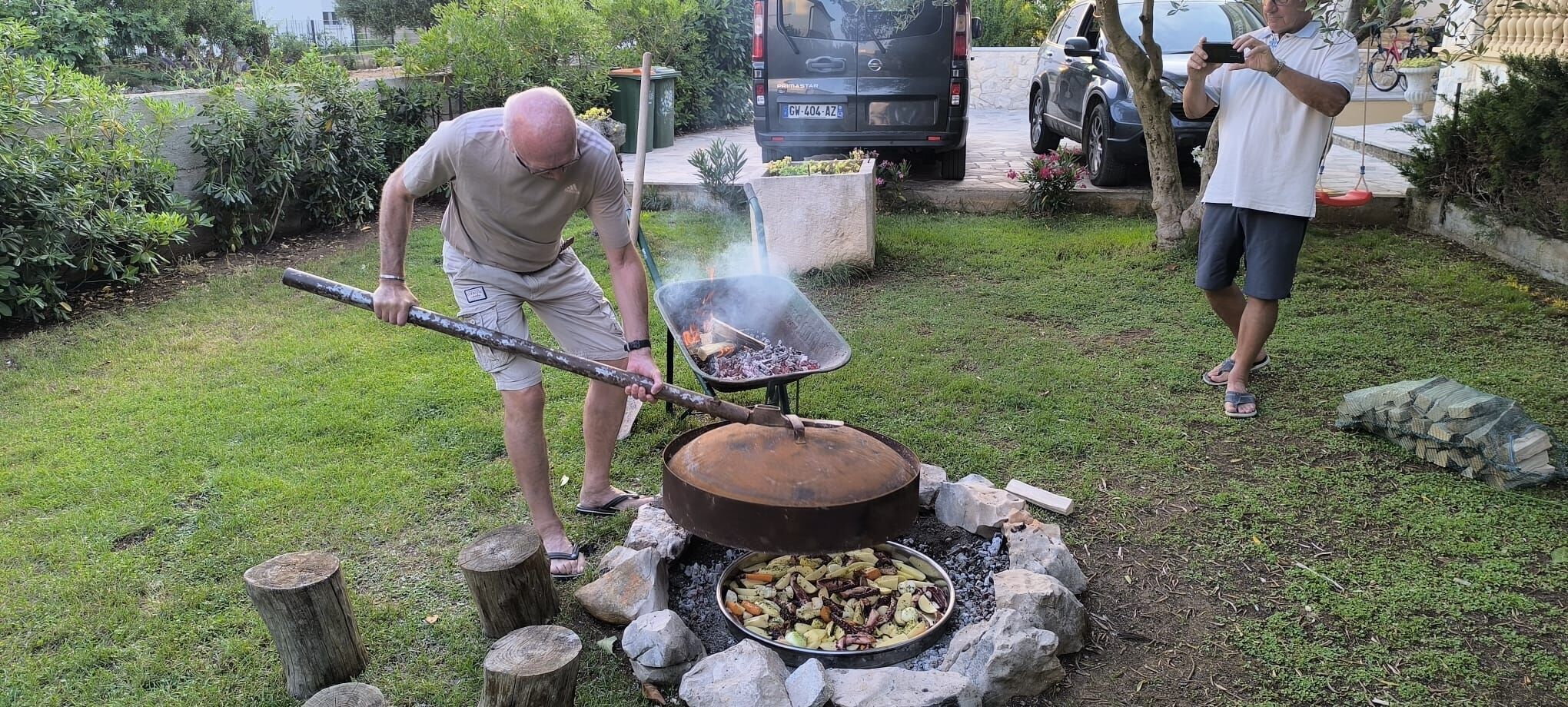 A man cooking outdoors with a large pot over an open fire, surrounded by rocks and logs.