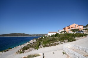 A coastal road with a stone retaining wall, a white building with a red roof, and a parked car.