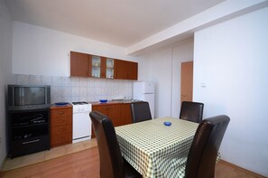 Μια μικρή, λειτουργική κουζίνα με γκαζιέρα, φούρνο μικροκυμάτων και λευκό ψυγείο. Υπάρχει ένα τραπέζι φαγητού με τέσσερις καρέκλες και ένα τραπεζομάντιλο με καρό σχέδιο.