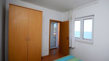 A bedroom with a large wardrobe, a bed with a striped bedspread, a window with a view of the sea, and a door leading to another room.