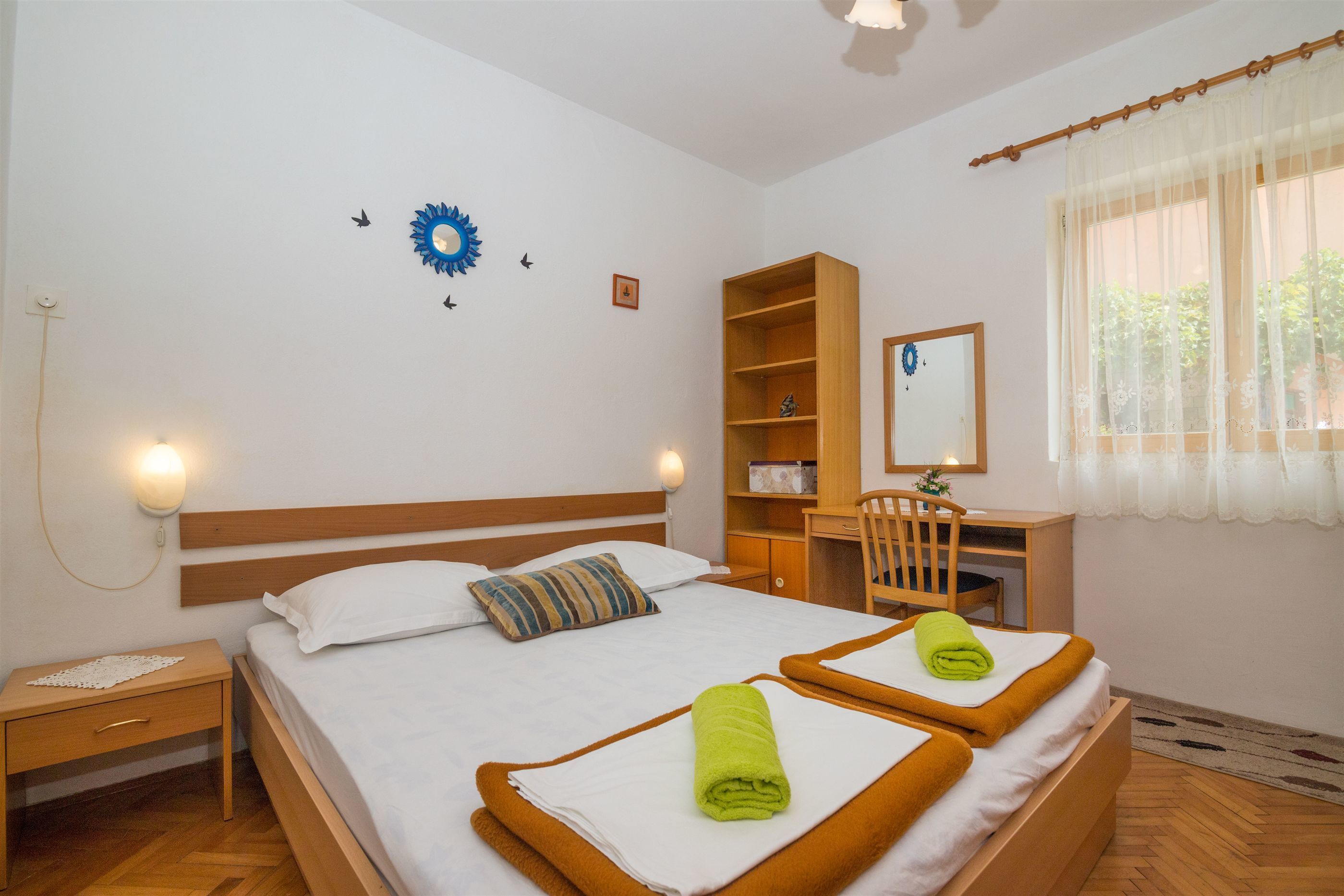 Une chambre d’hôtel avec deux lits, une table de chevet en bois, un bureau, une chaise, un miroir et une fenêtre avec des rideaux légers.