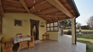 Un espacio cubierto con mesa y bancos de madera, una ventana pequeña y vistas al jardín.