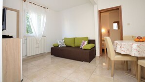 Una sala de estar con un sofá marrón, una mesa de comedor con mantel floreado y una ventana con cortinas blancas.