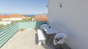 A balcony with a white table and bench, a green and white striped barrier, and a view of buildings and mountains in the distance. - House Jadranka (Supetar)